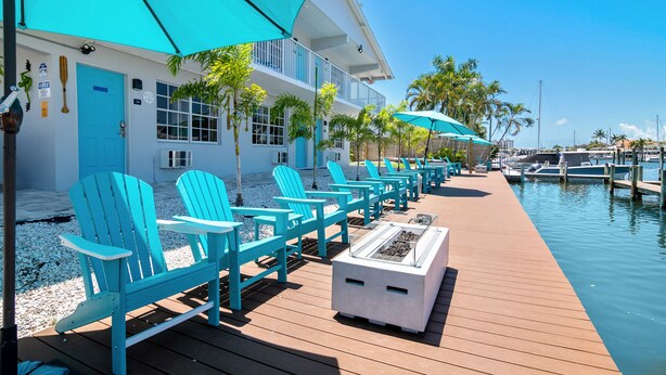 A row of turquoise Adirondack chairs with umbrellas, a wooden deck, and a marina in the background. - Latitude 26 Waterfront Boutique Resort - Fort Myers Beach (Fort Myers Beach)