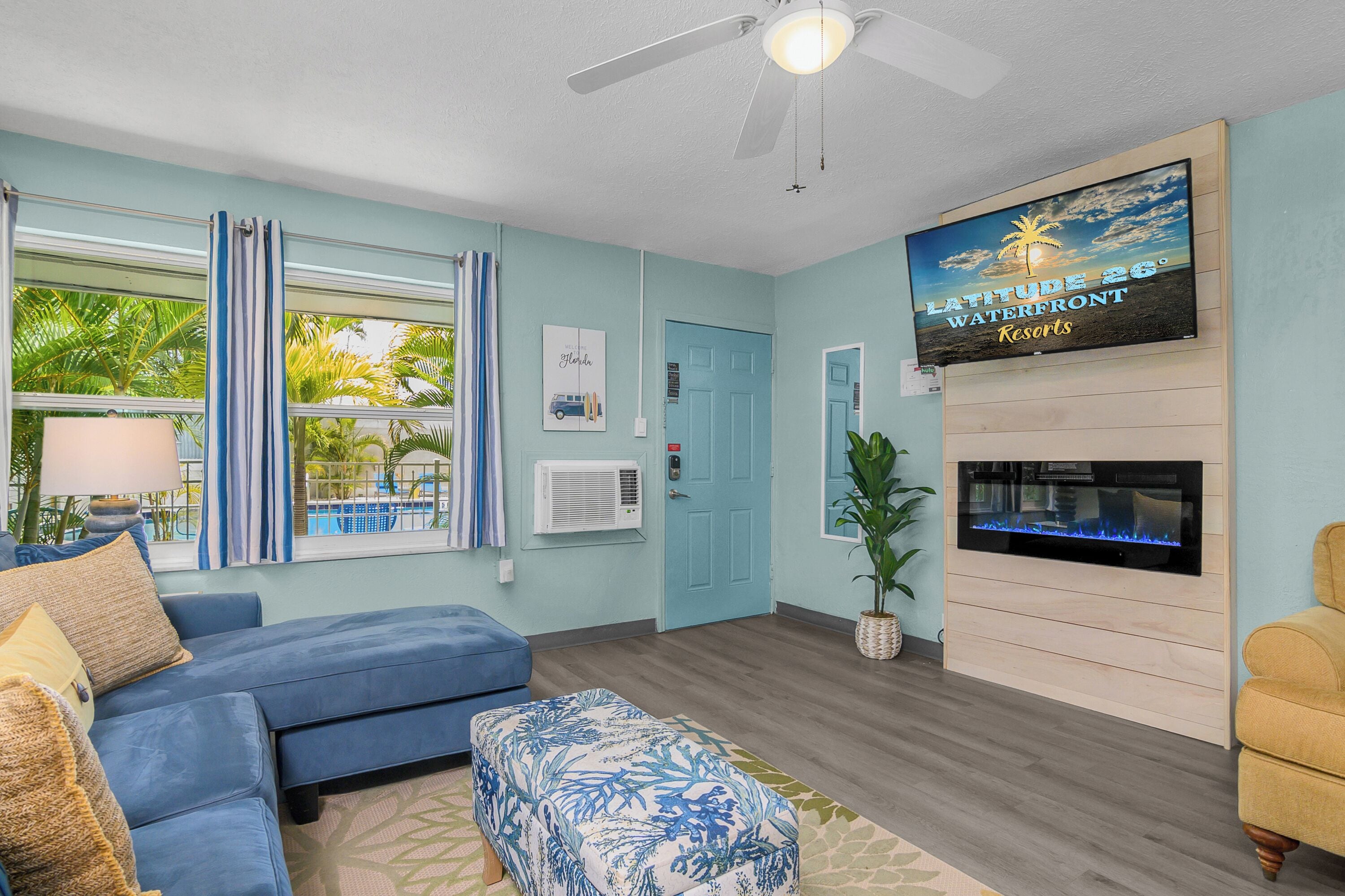 Una sala de estar con un sofá azul, un otoman estampado, un televisor de pantalla plana, un ventilador de techo y un letrero de Latitude 26° Waterfront Resorts.