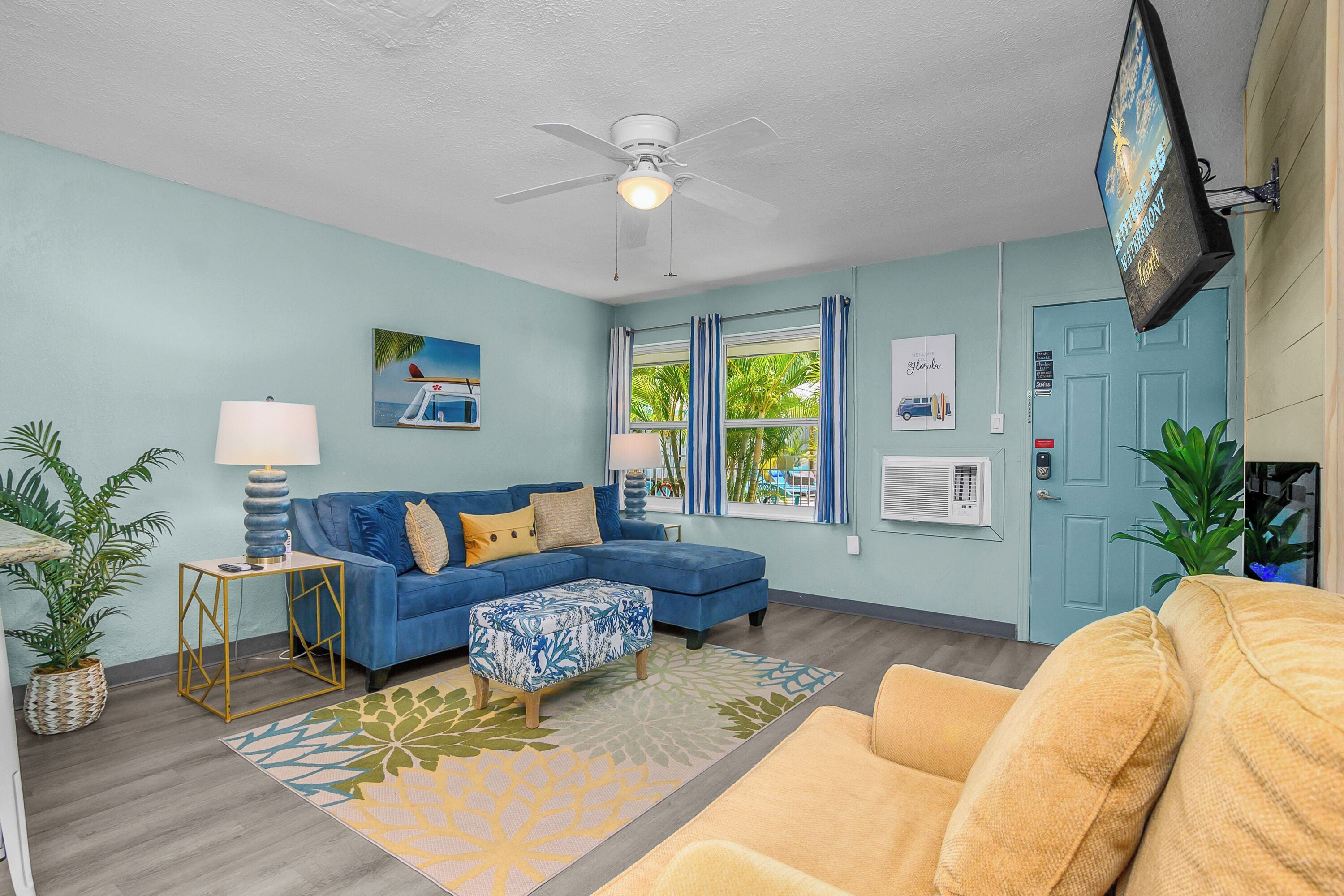 A living room with a blue sofa, a yellow sofa, a ceiling fan, a TV mounted on the wall, and a door with an air conditioning unit.