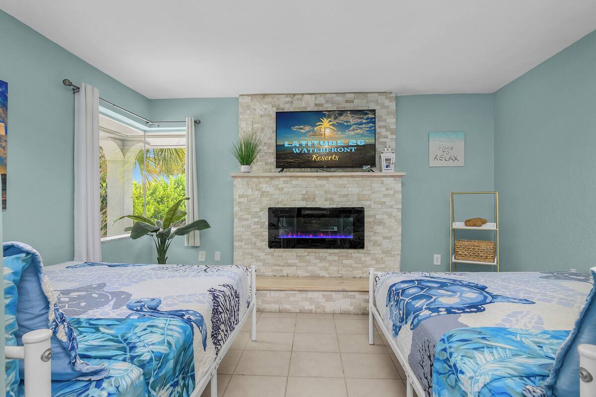 A bedroom with two beds, a TV mounted above a fireplace, and a view of greenery outside.