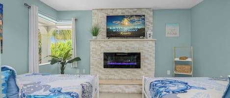 A bedroom with two beds, a TV mounted above a fireplace, and a view of greenery outside.