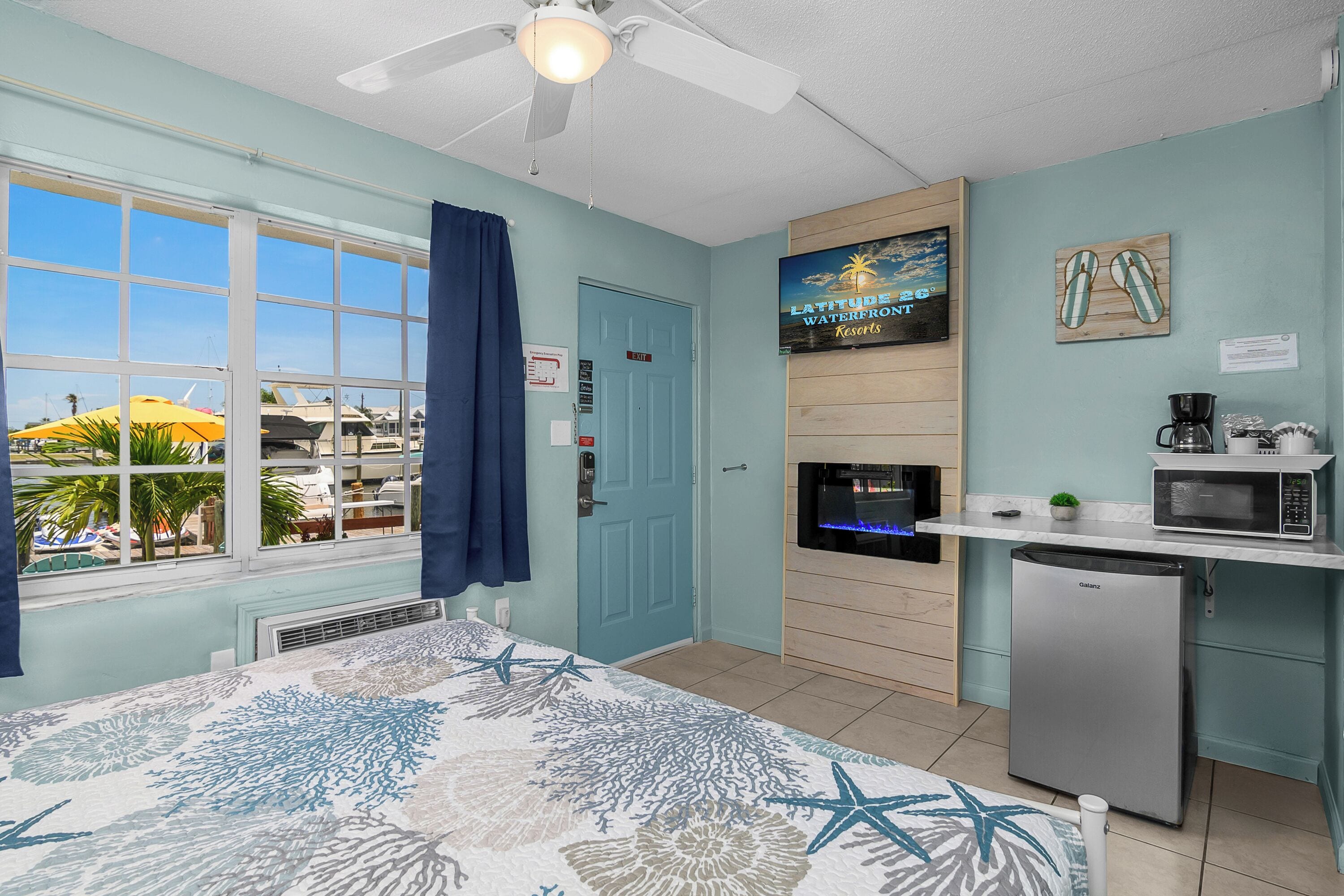 A room with a bed, a ceiling fan, a kitchenette with a refrigerator, a microwave, and a coffee maker. There is a door with a sign, a window with curtains, and a view of palm trees and boats outside.