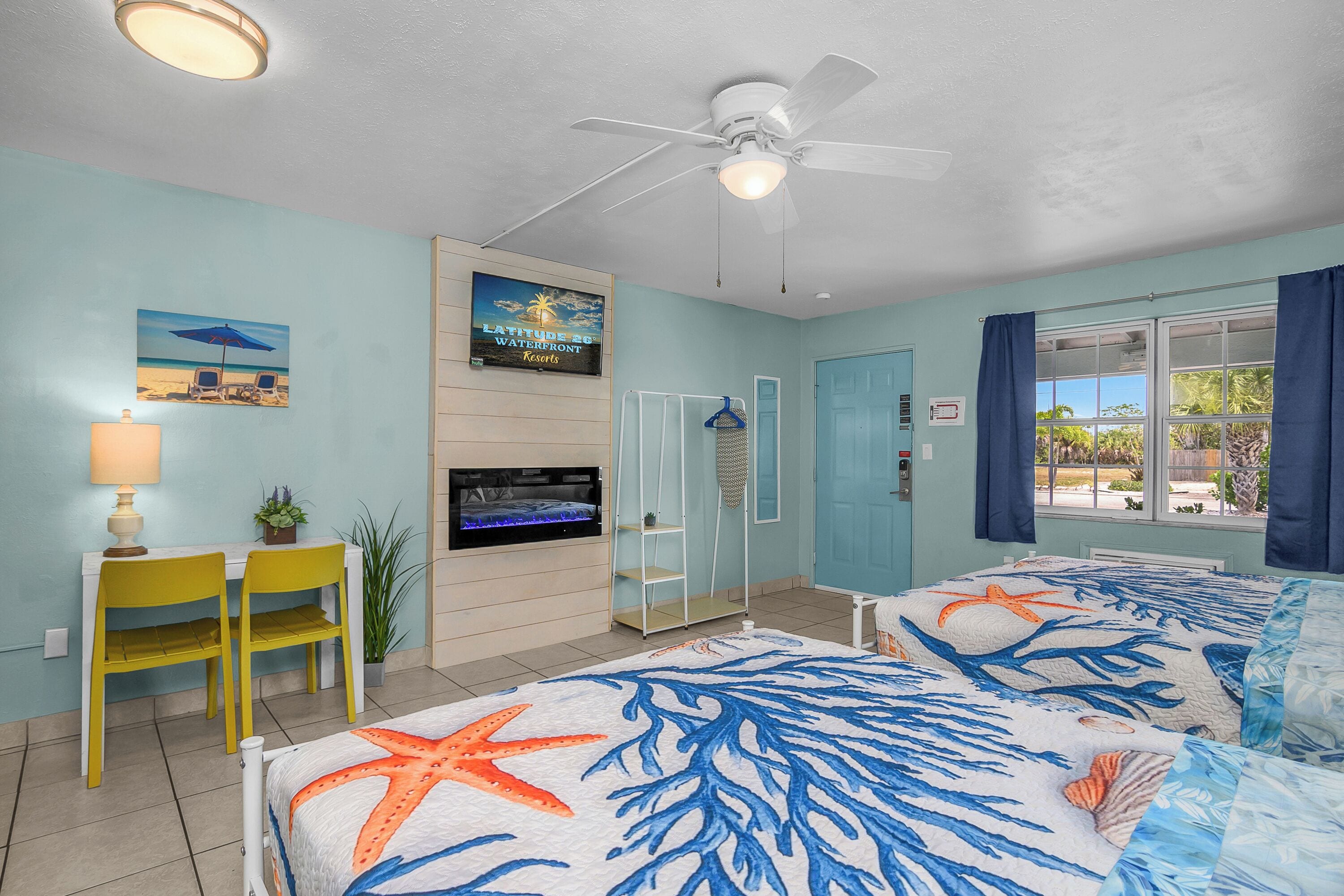 A room with two beds, a ceiling fan, a TV, a desk with chairs, a plant, and a beach-themed wall art.