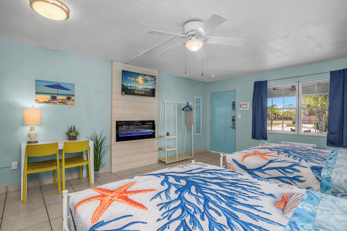A room with two beds, a ceiling fan, a TV, a desk with chairs, a plant, and a beach-themed wall art.