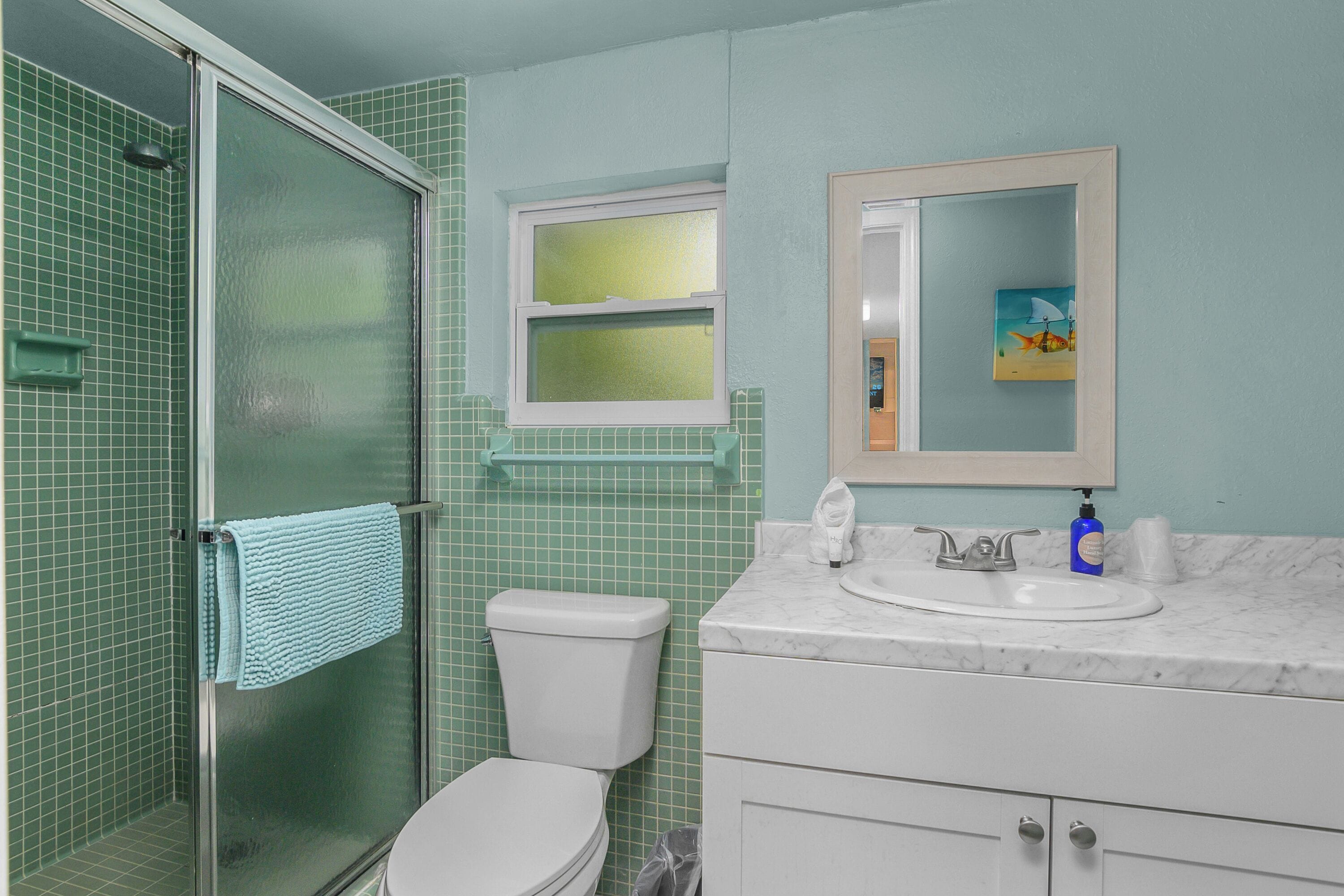 A bathroom with a glass shower enclosure, a toilet, a sink with a marble countertop, and a mirror with a framed artwork.