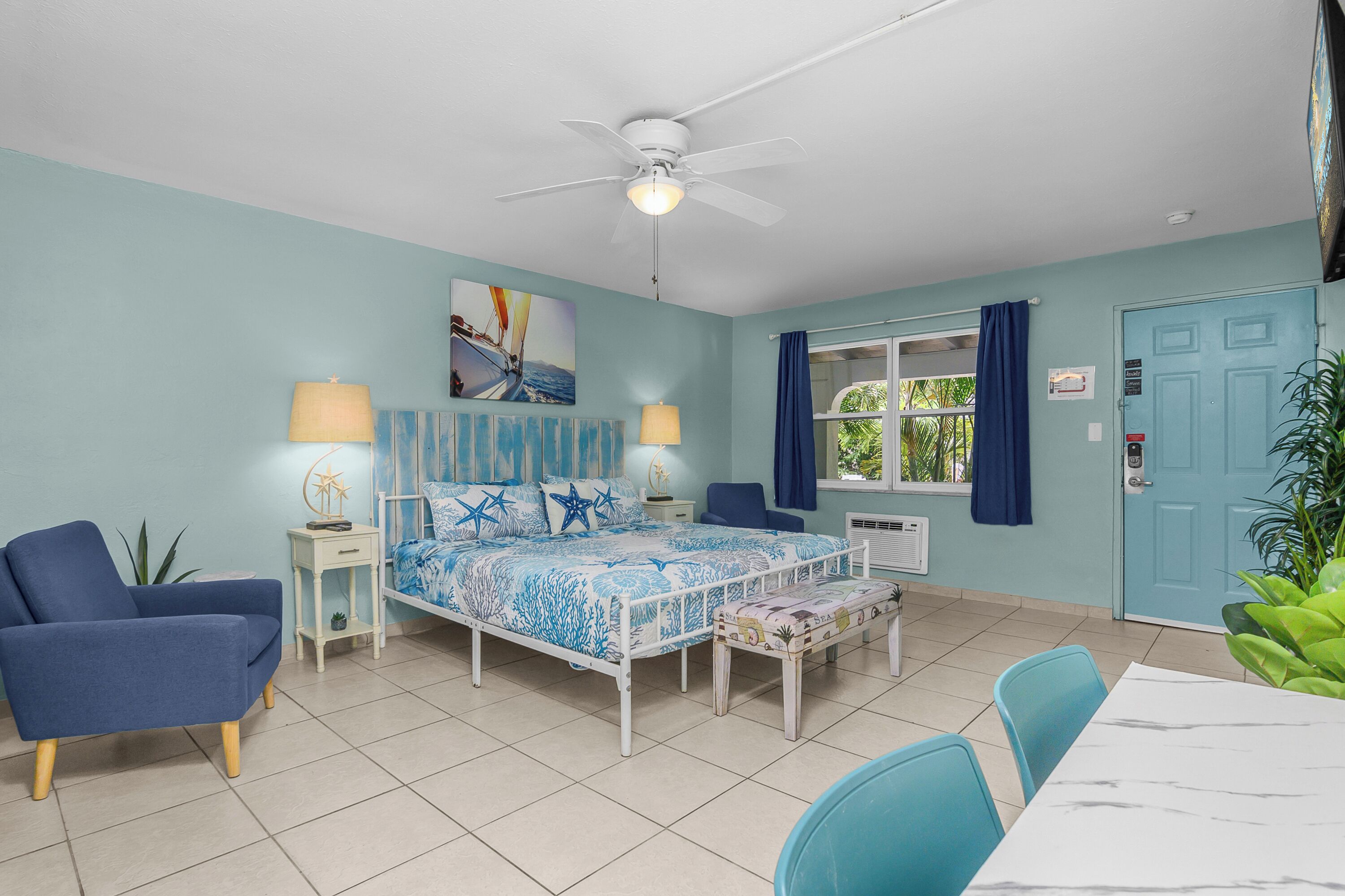 A bedroom with a bed, bedside tables, a chair, a table, a ceiling fan, a window with curtains, and a door.