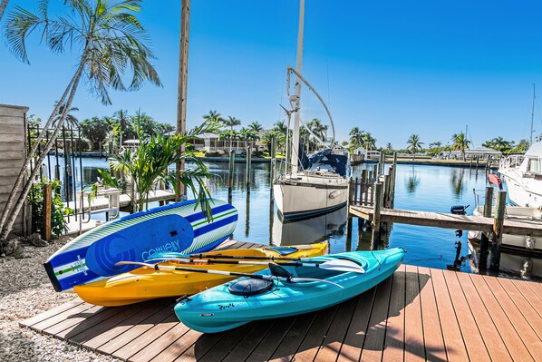 Un muelle de madera con kayaks y una lancha amarrados en un puerto deportivo, con agua azul cristalina y palmeras al fondo.