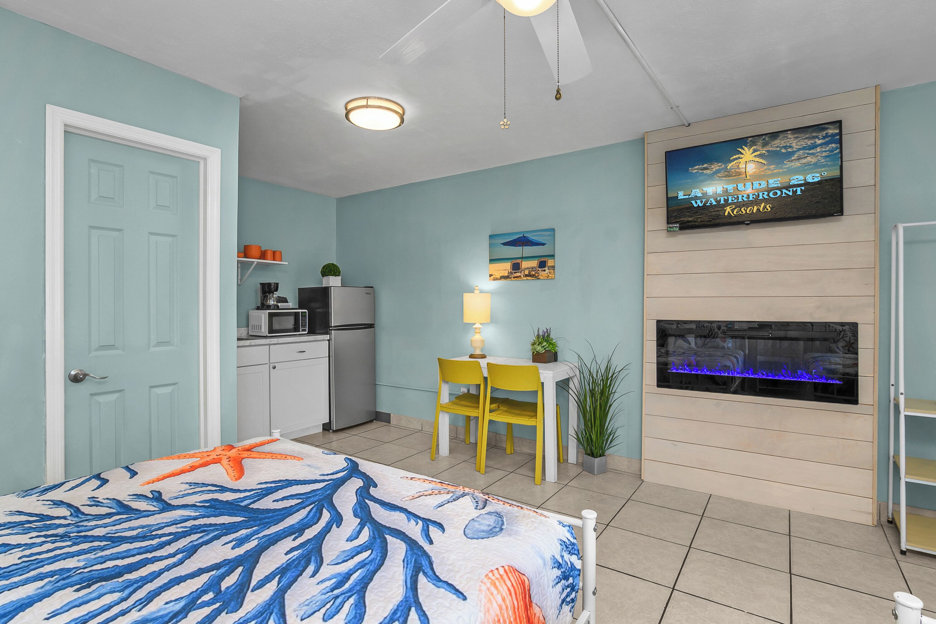 A room with a bed, a TV mounted on the wall, a small kitchenette, a dining area with chairs, and a fireplace.