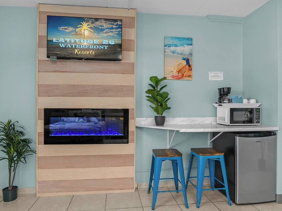 A kitchen with a flat-screen TV, a microwave, a refrigerator, and two blue stools.