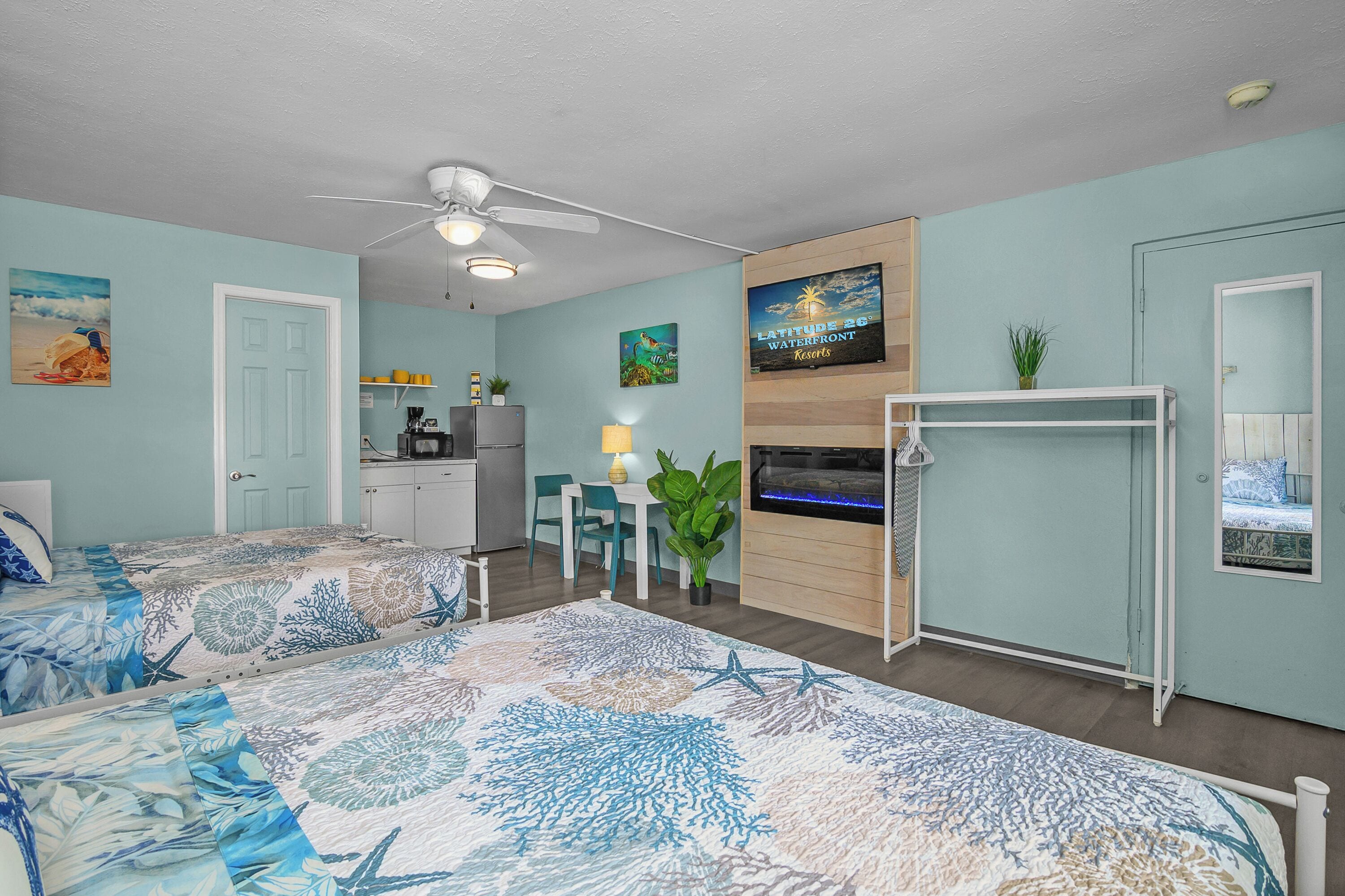 A room with two beds, a ceiling fan, a kitchenette, a dining area, and a TV displaying a resort sign.