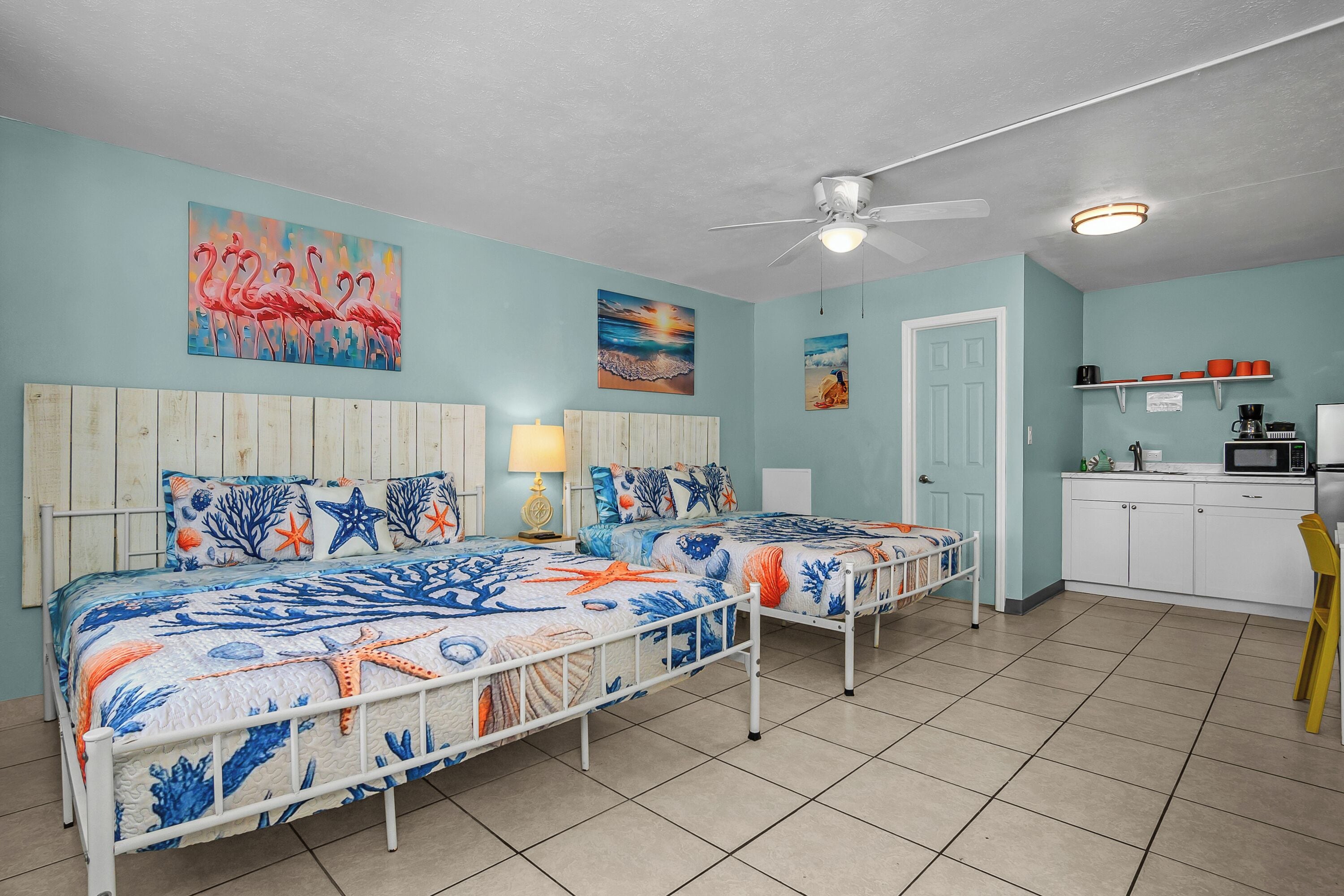 Une chambre avec deux lits, un ventilateur de plafond, un coin cuisine et des œuvres d’art accrochées aux murs.