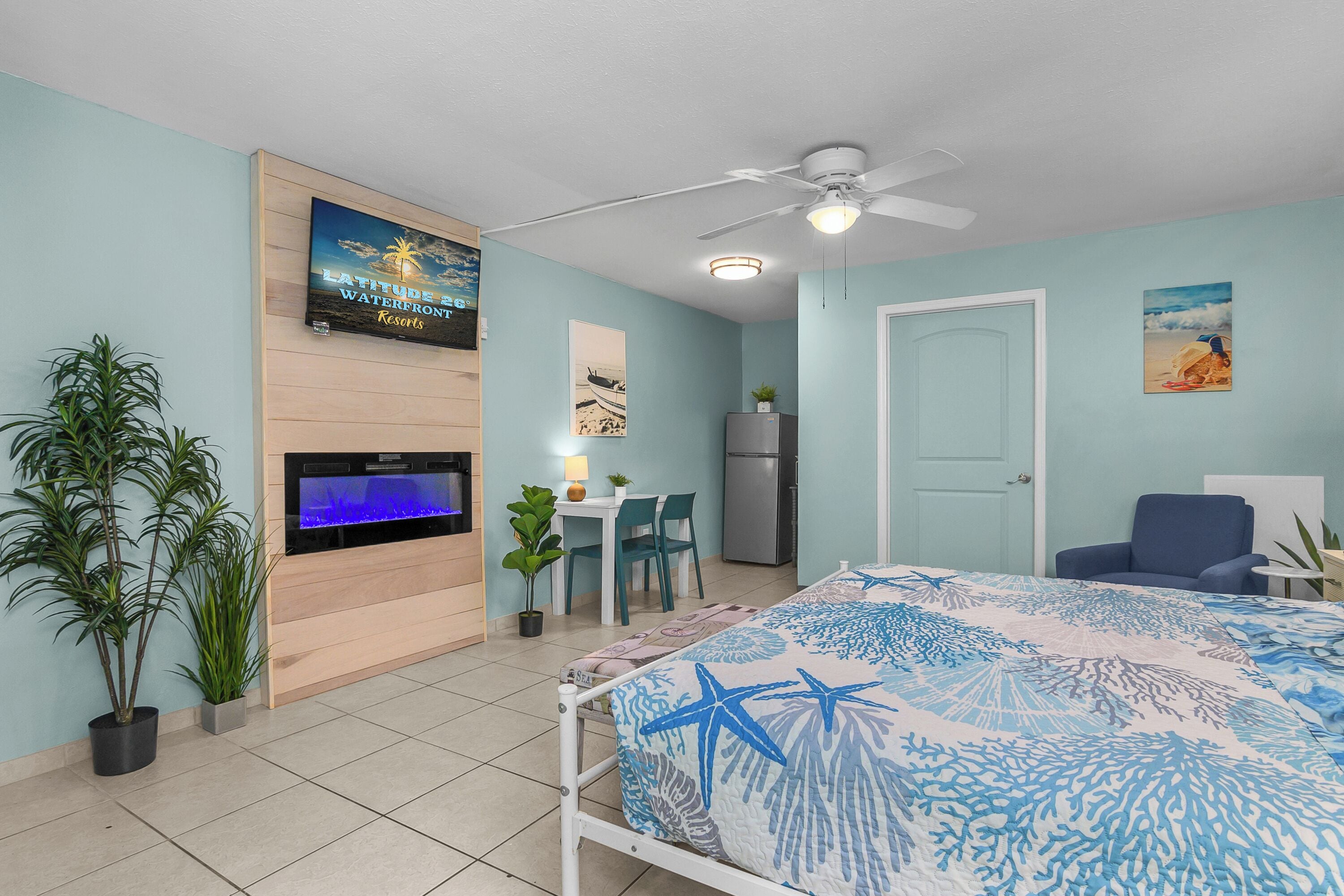 Un dormitorio con cama, ventilador de techo, televisor, escritorio con silla, refrigerador y una puerta.