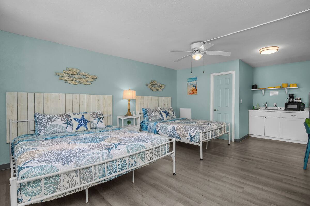 A bedroom with two beds, a ceiling fan, a nightstand with a lamp, and a kitchen area with a microwave.