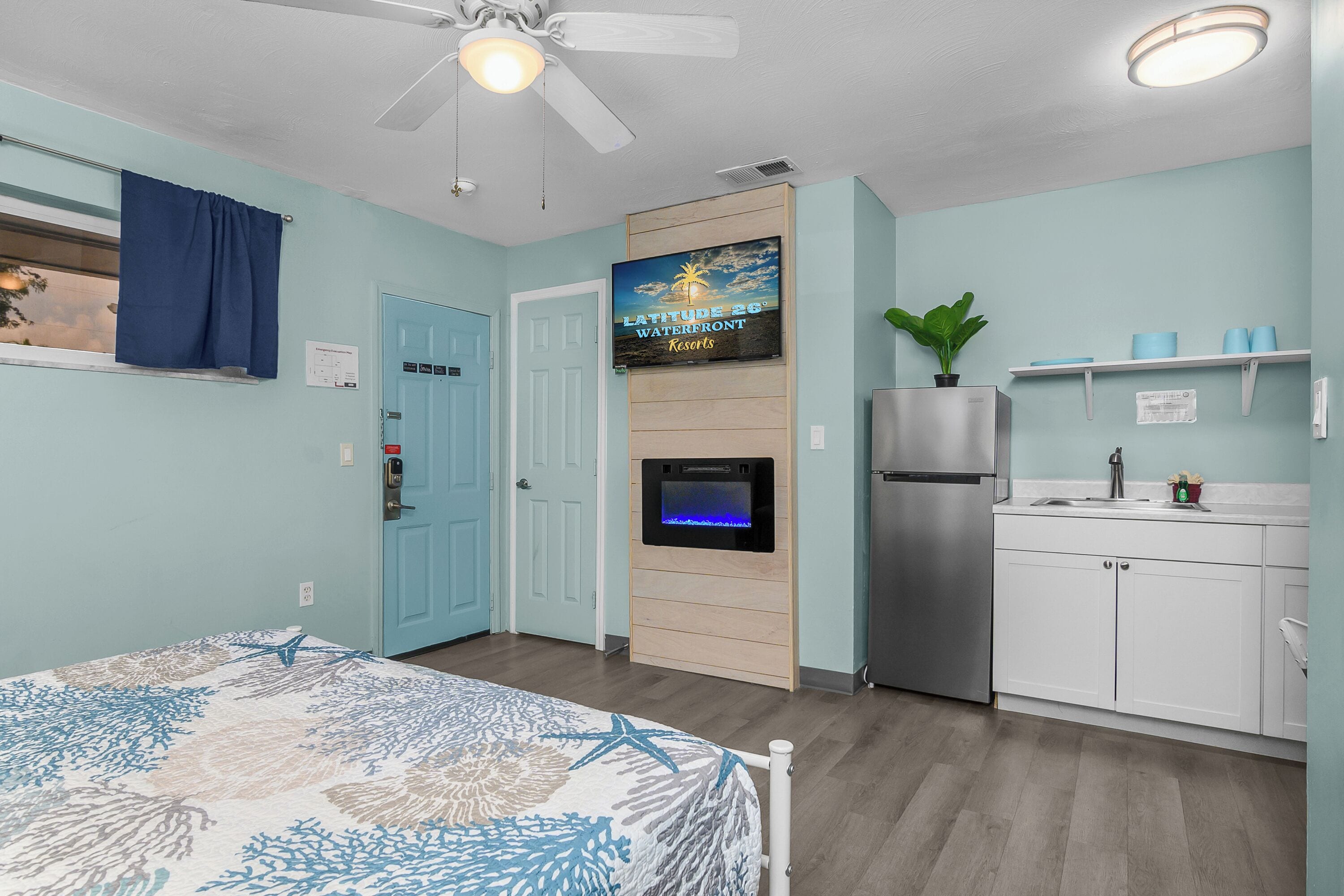 A room with a bed, a ceiling fan, a TV mounted on a wooden panel, a refrigerator, a sink, and a window with a curtain.