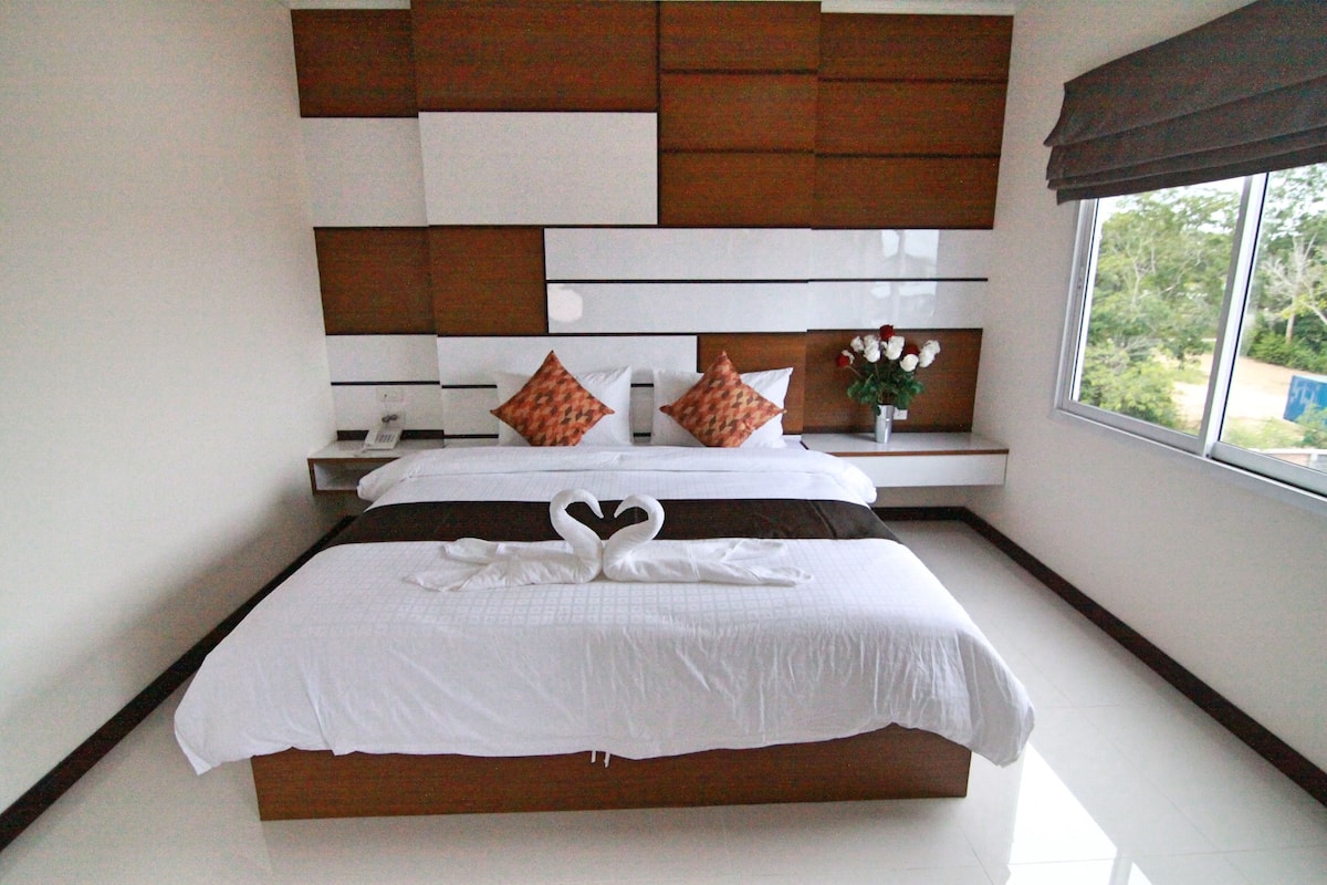 A neatly made bed with a heart-shaped towel arrangement, a bedside table with a vase of flowers, and a view of greenery through a window.