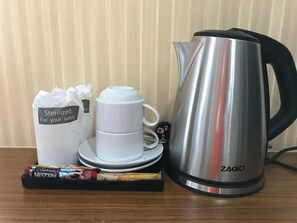 A stainless steel ZAGIO electric kettle, a white coffee cup and saucer, a white bag labeled "Sterilized For your safety", and a Nescafe coffee packet on a wooden table. - TTT Hotel (Hat Yai)