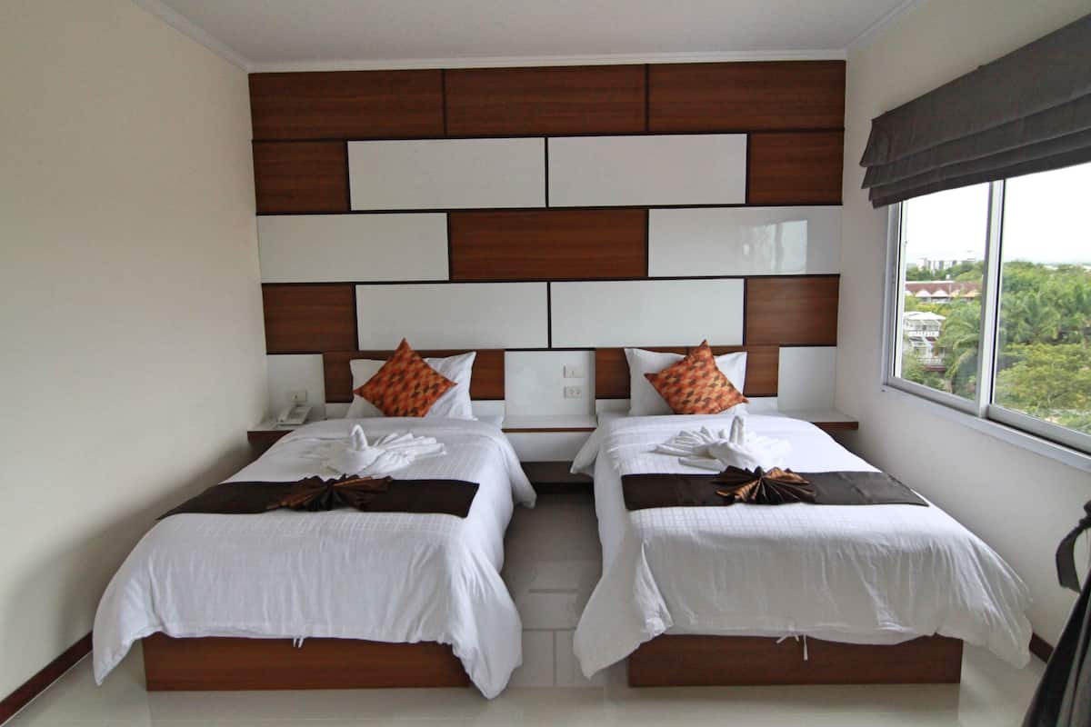 A hotel room with two beds, a wooden headboard, a large window, and a view of greenery outside.