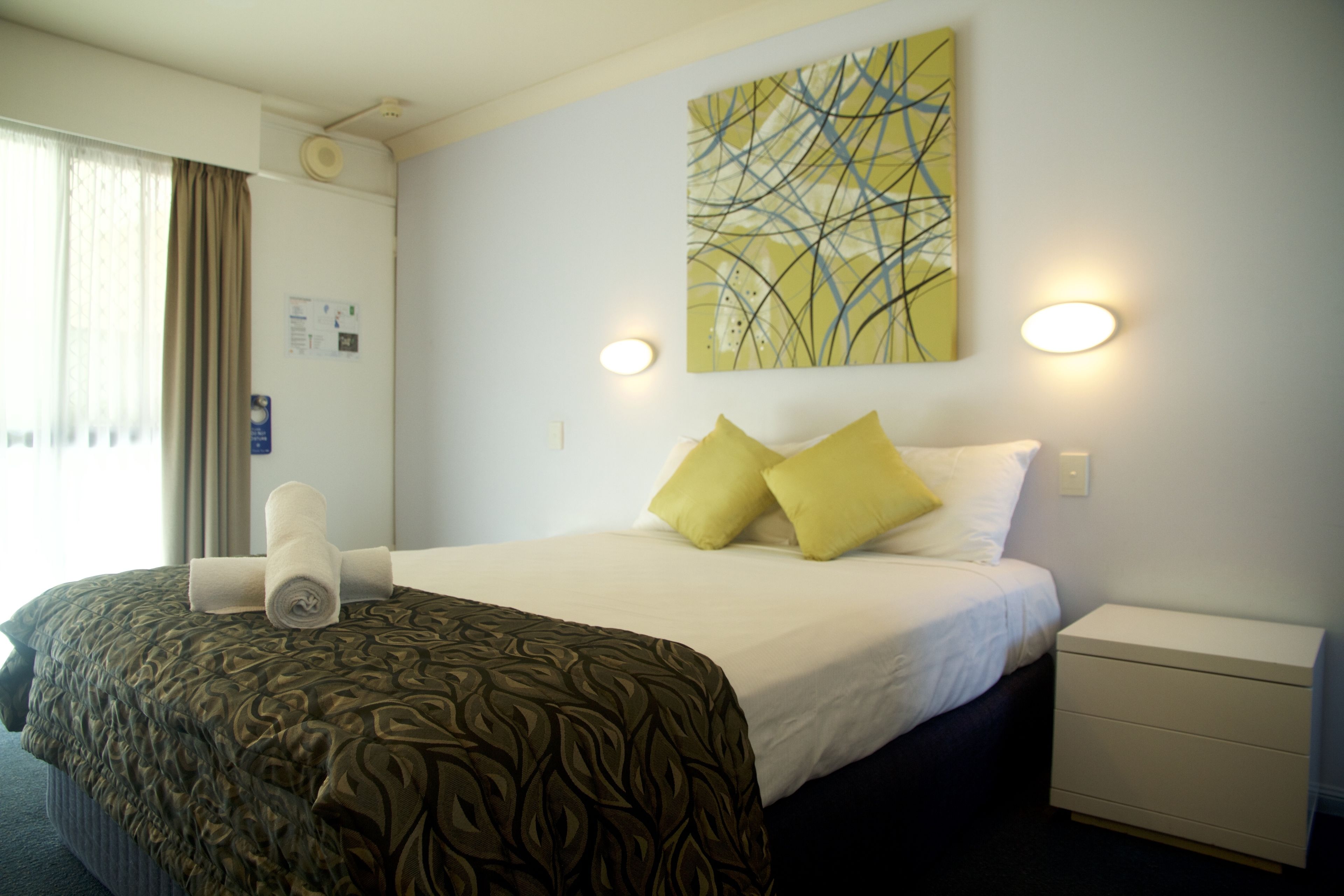 A hotel room with a neatly made bed, a bedside table, a wall-mounted artwork, and a window with curtains.