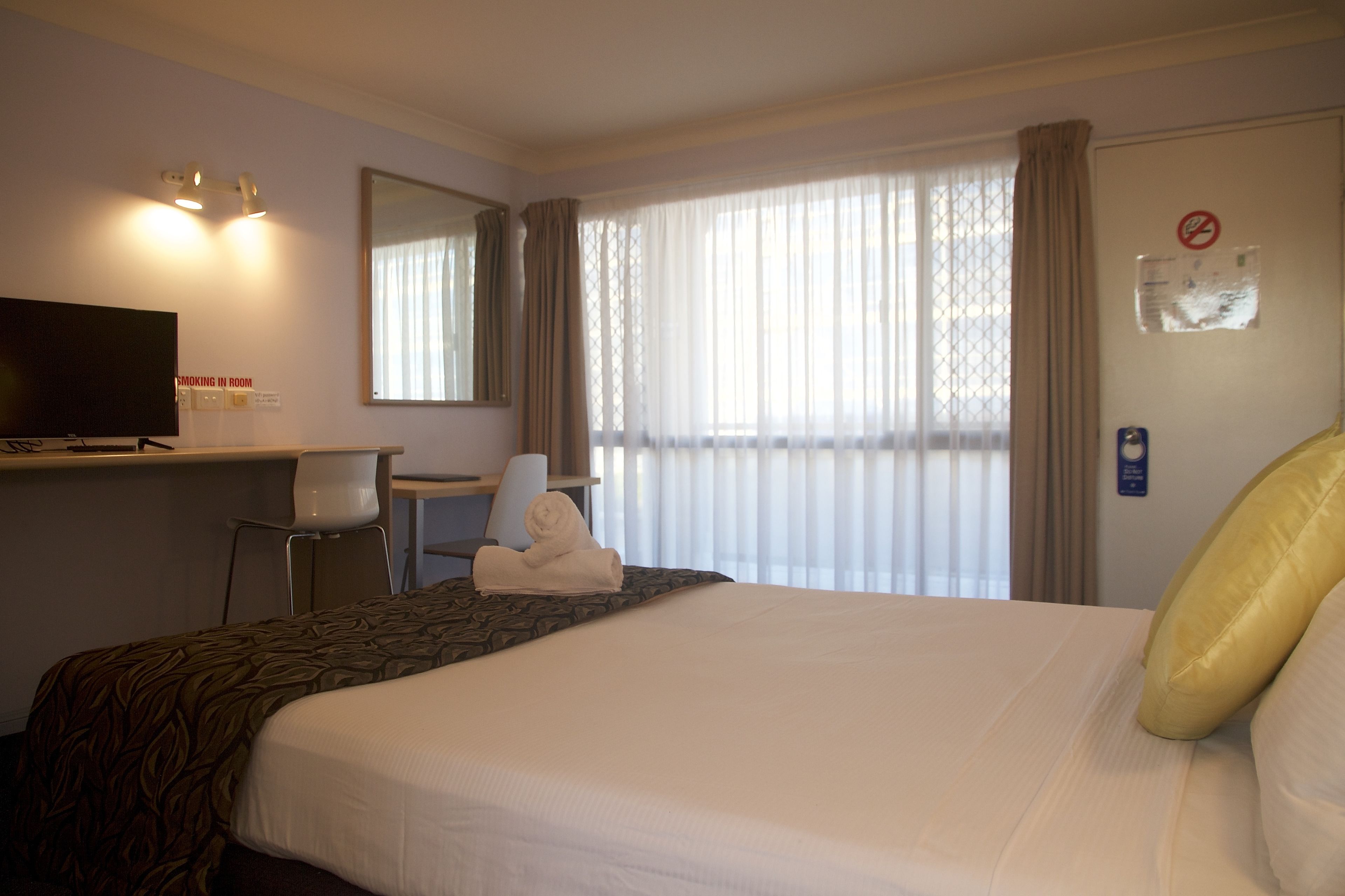 A hotel room with a bed, a desk with a television, a chair, and a window with curtains.