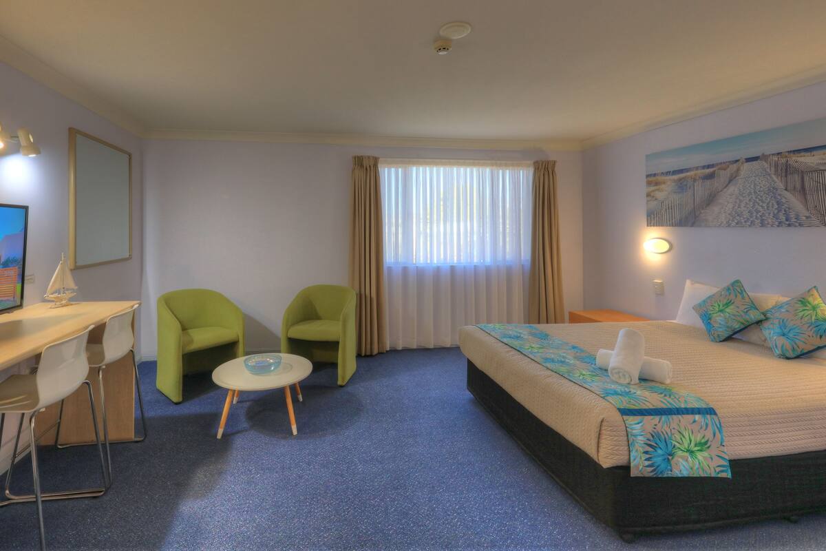 A hotel room with a bed, two chairs, a desk with a TV, and a window with curtains.