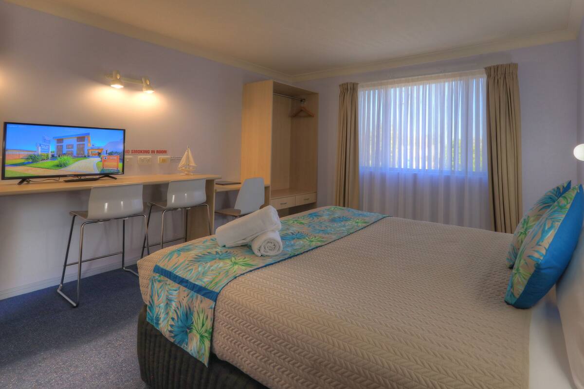 A hotel room with a bed, a desk with a chair, a television, and a window with curtains.