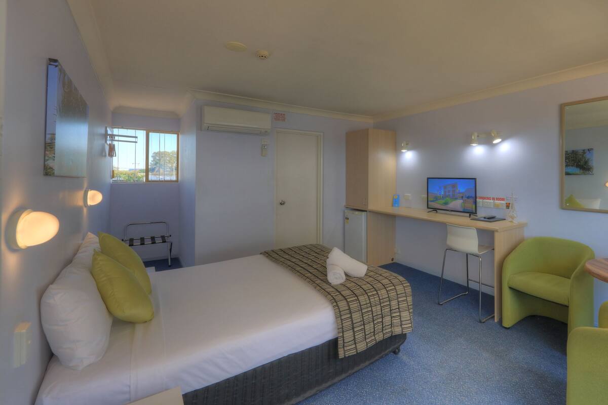 A hotel room with a bed, a desk, a chair, a small table, a mirror, and a television.