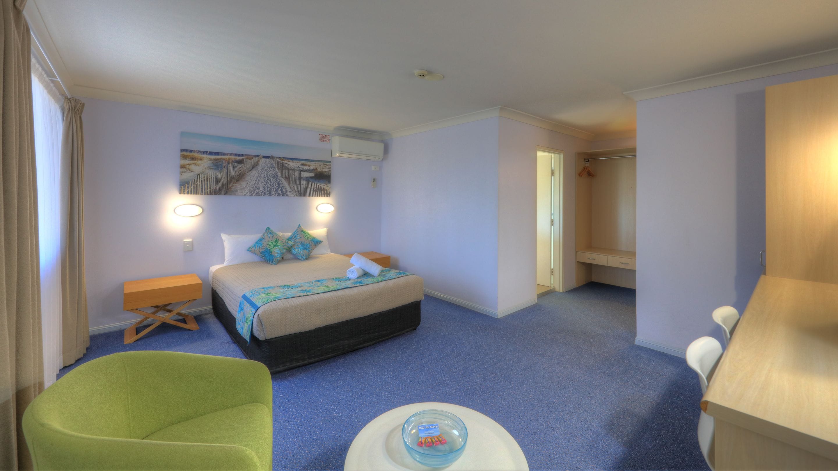 Una habitación de hotel con cama, una silla verde, una mesita con un cuenco y un armario.