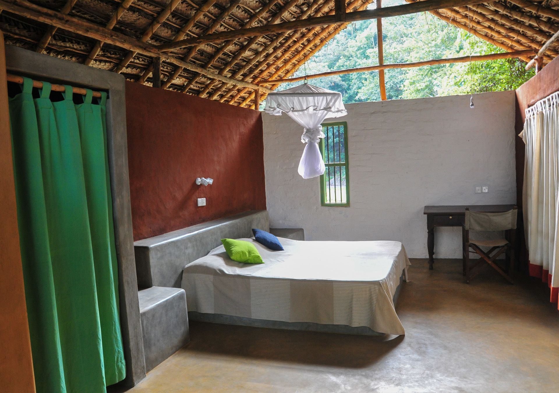 A room with a bed, a desk, a chair, a hanging mosquito net, and a window with a green frame.