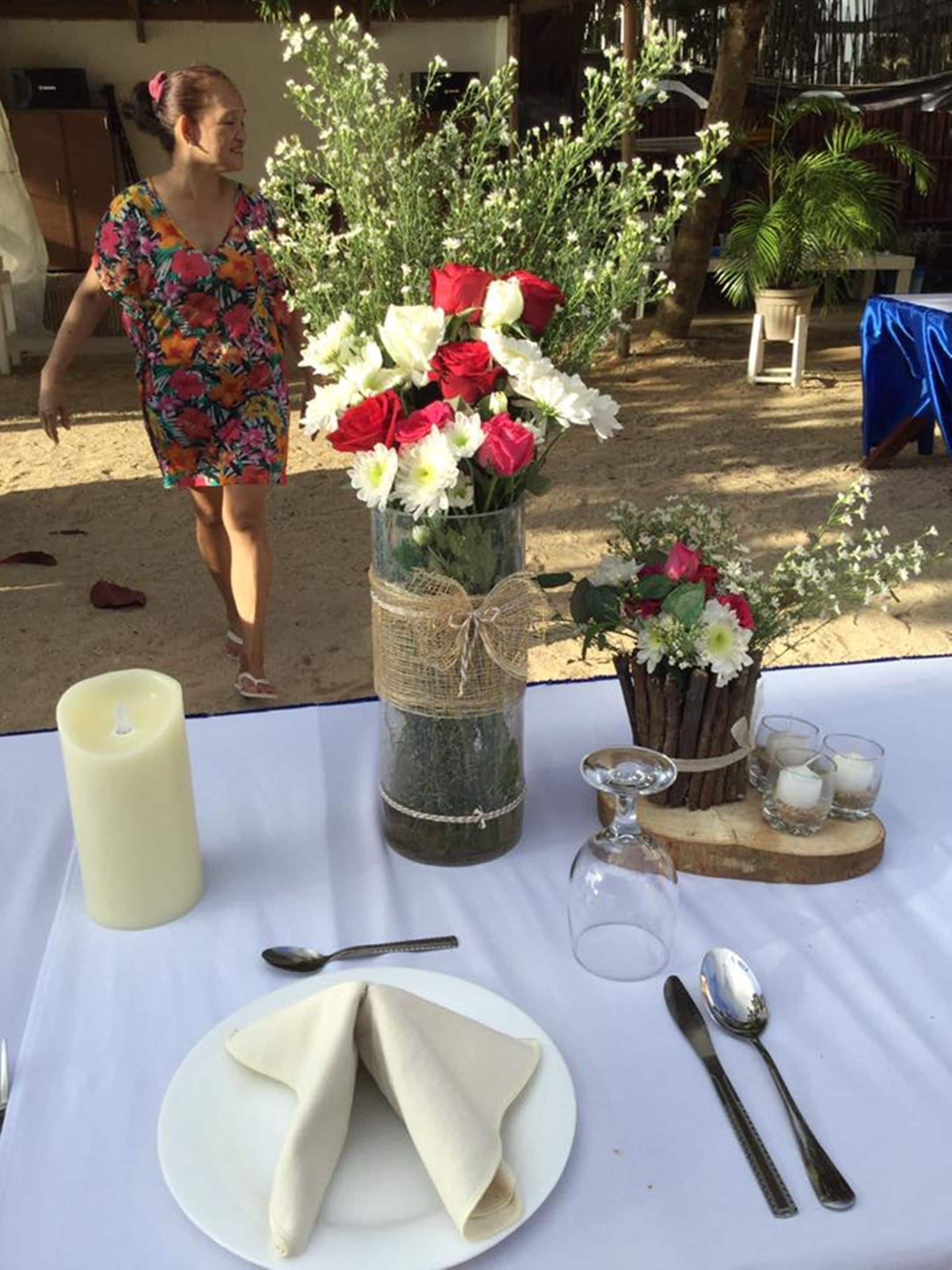 A table set outdoors with a white cloth, a candle, a vase of flowers, and cutlery.