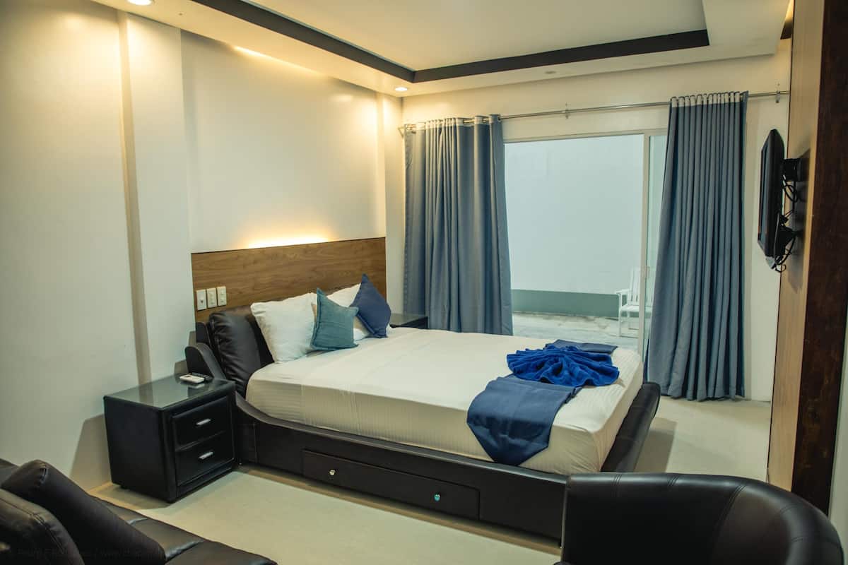 A modern bedroom with a large bed, a nightstand, a chair, and a view of the outdoors through a window with curtains.