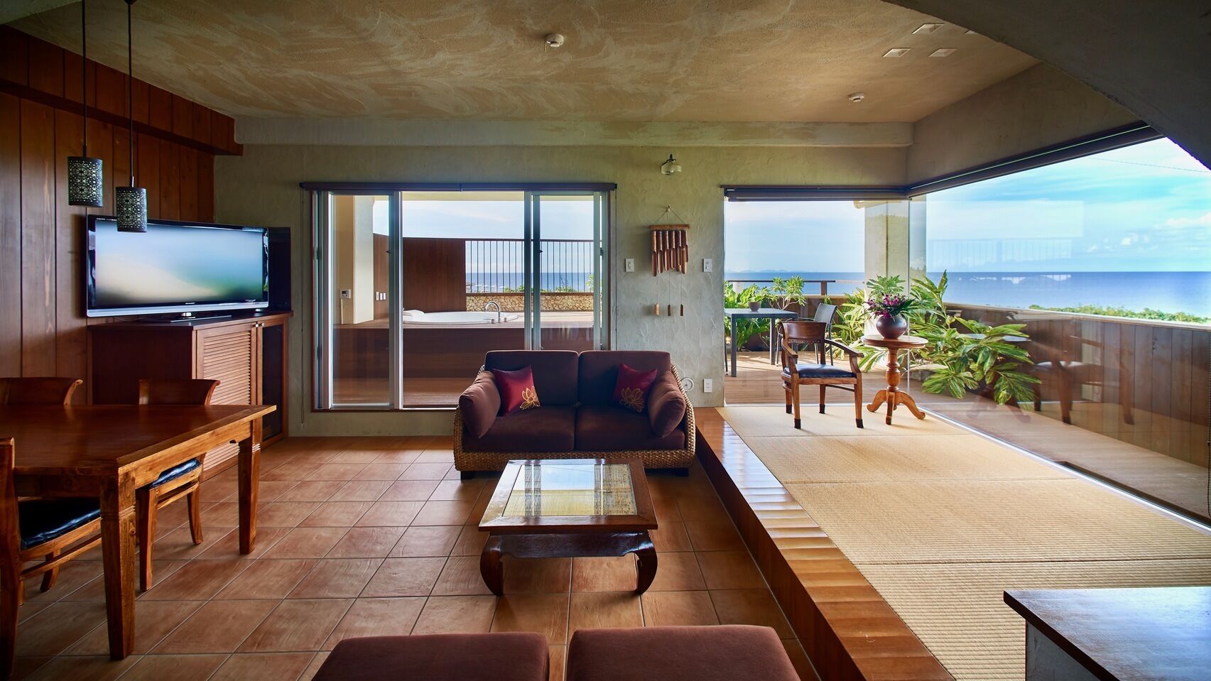 A spacious living area with a large TV, a sofa, a dining table, and a balcony with a view of the ocean.