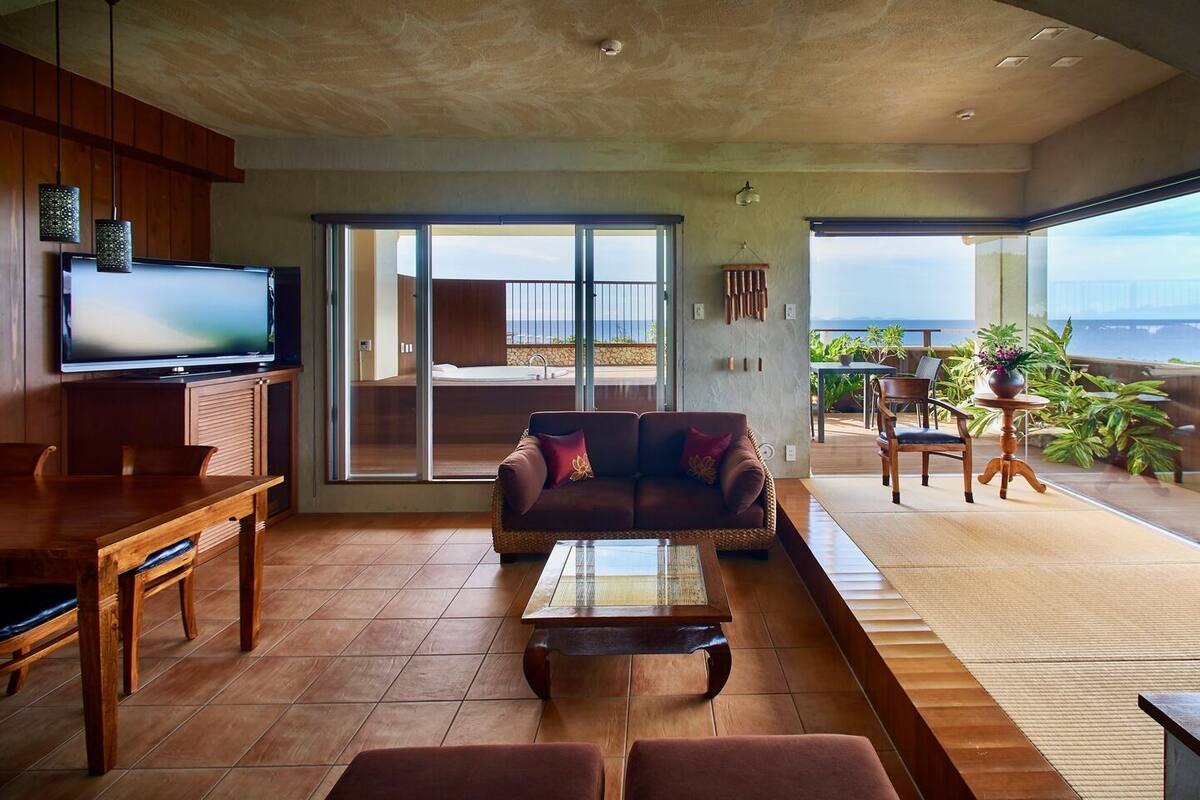 Amplia sala de estar con televisor grande, sofá, mesa de comedor y balcón con vista al océano.