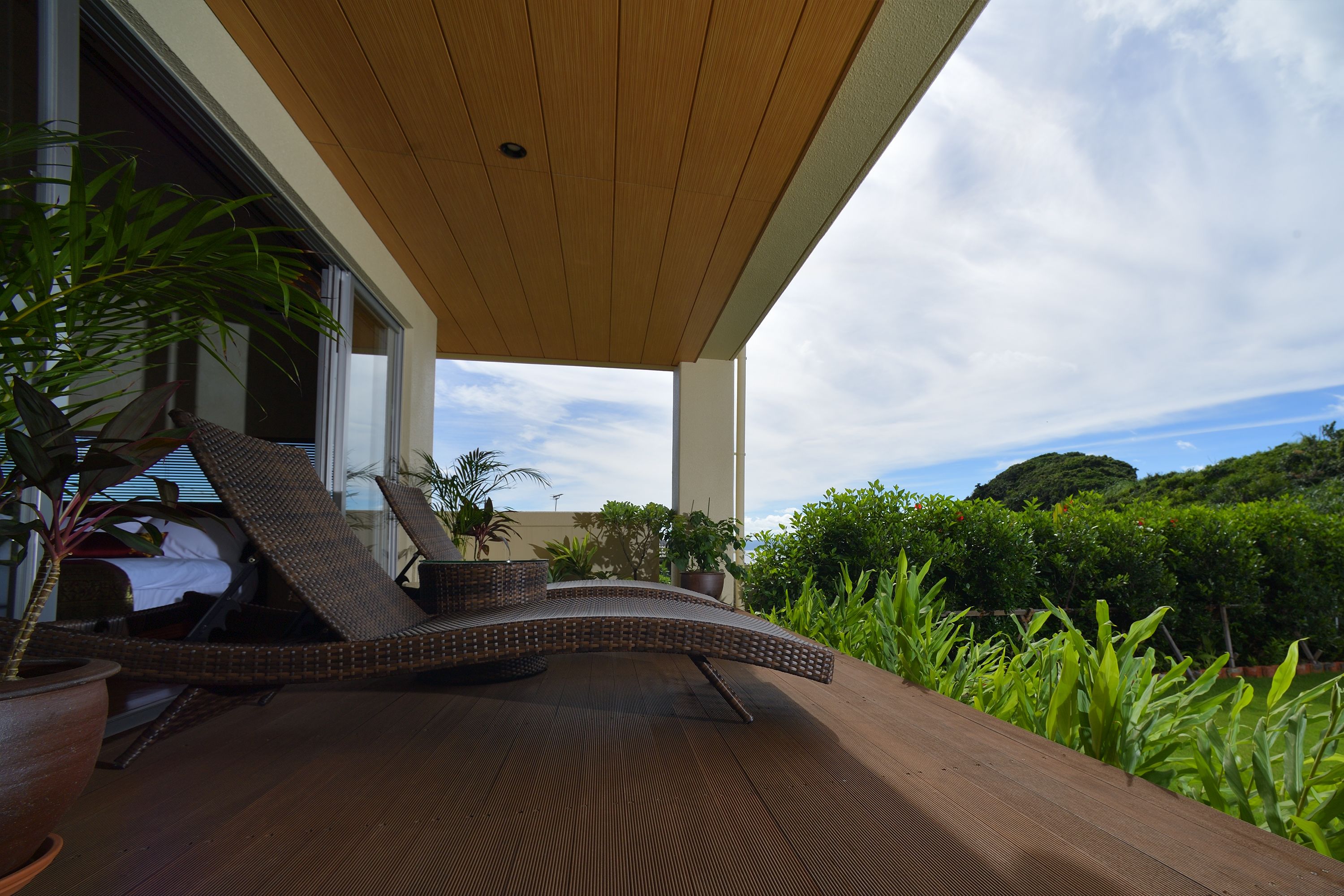 A wooden deck with a curved lounge chair, potted plants, and a view of lush greenery and hills.