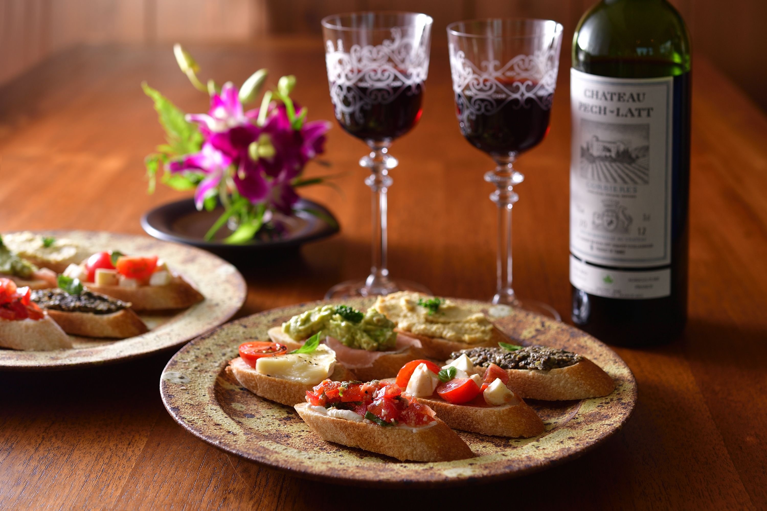 Two plates of bruschetta with various toppings, two wine glasses, and a bottle of Chateau Foch-Latt wine on a wooden table.