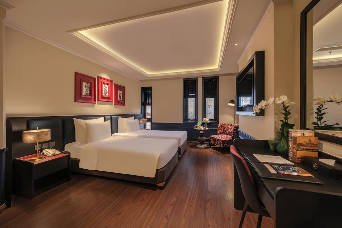 A hotel room with a large bed, two bedside tables, a desk, and a chair. There are framed pictures on the wall, a TV, and a window with blinds.