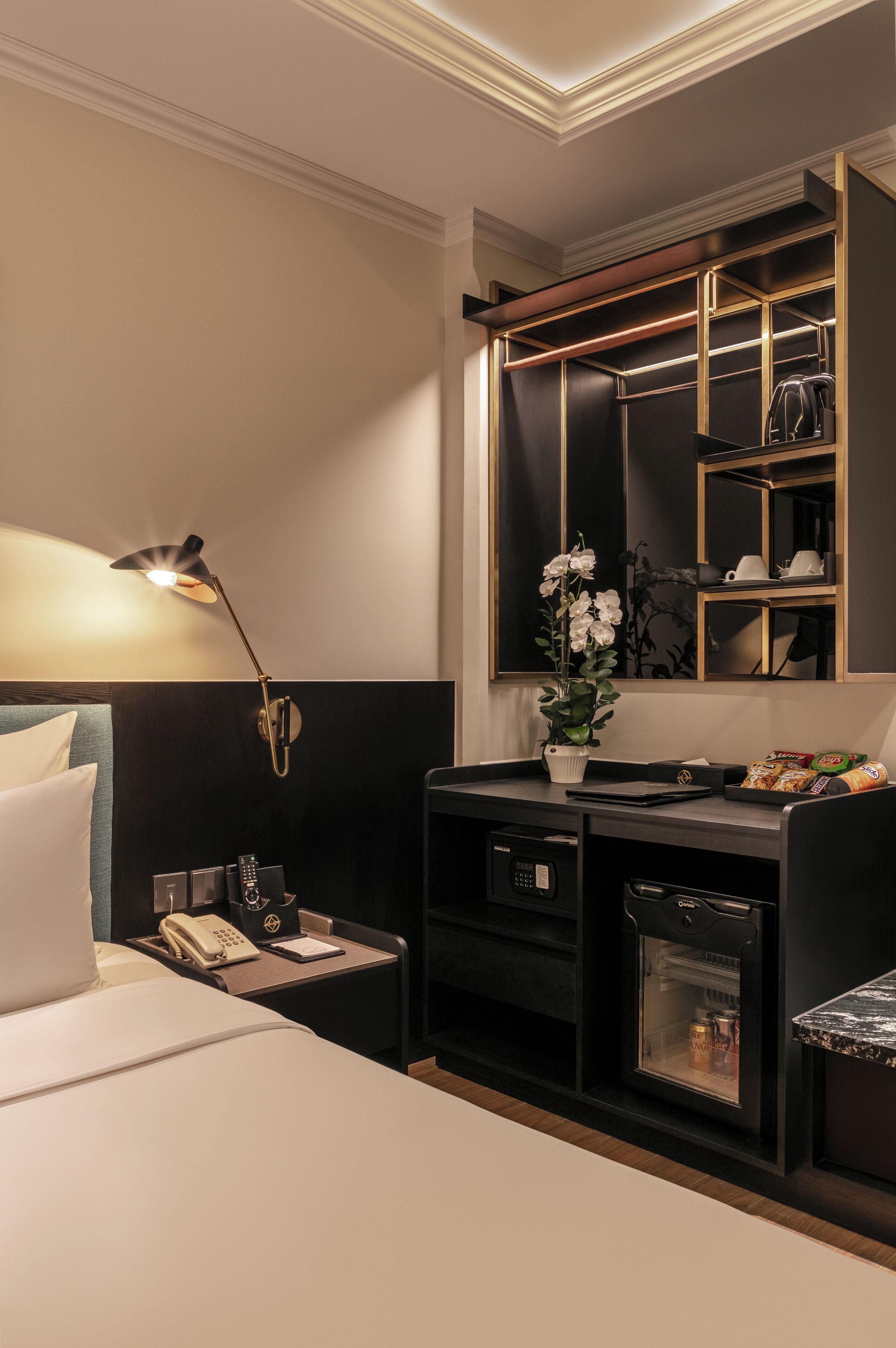 A hotel room with a bed, bedside table, lamp, minibar, and a mirror with shelves.