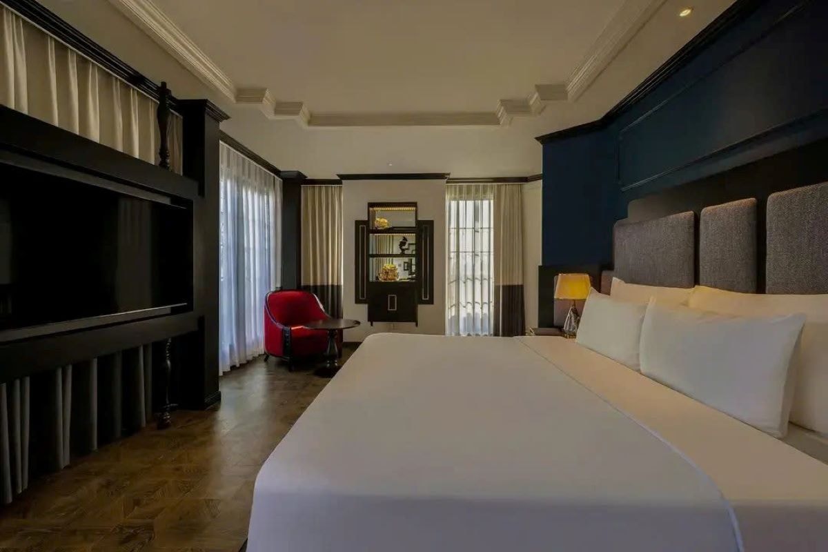 A hotel room with a large bed, a red chair, a television, and a nightstand with a lamp.
