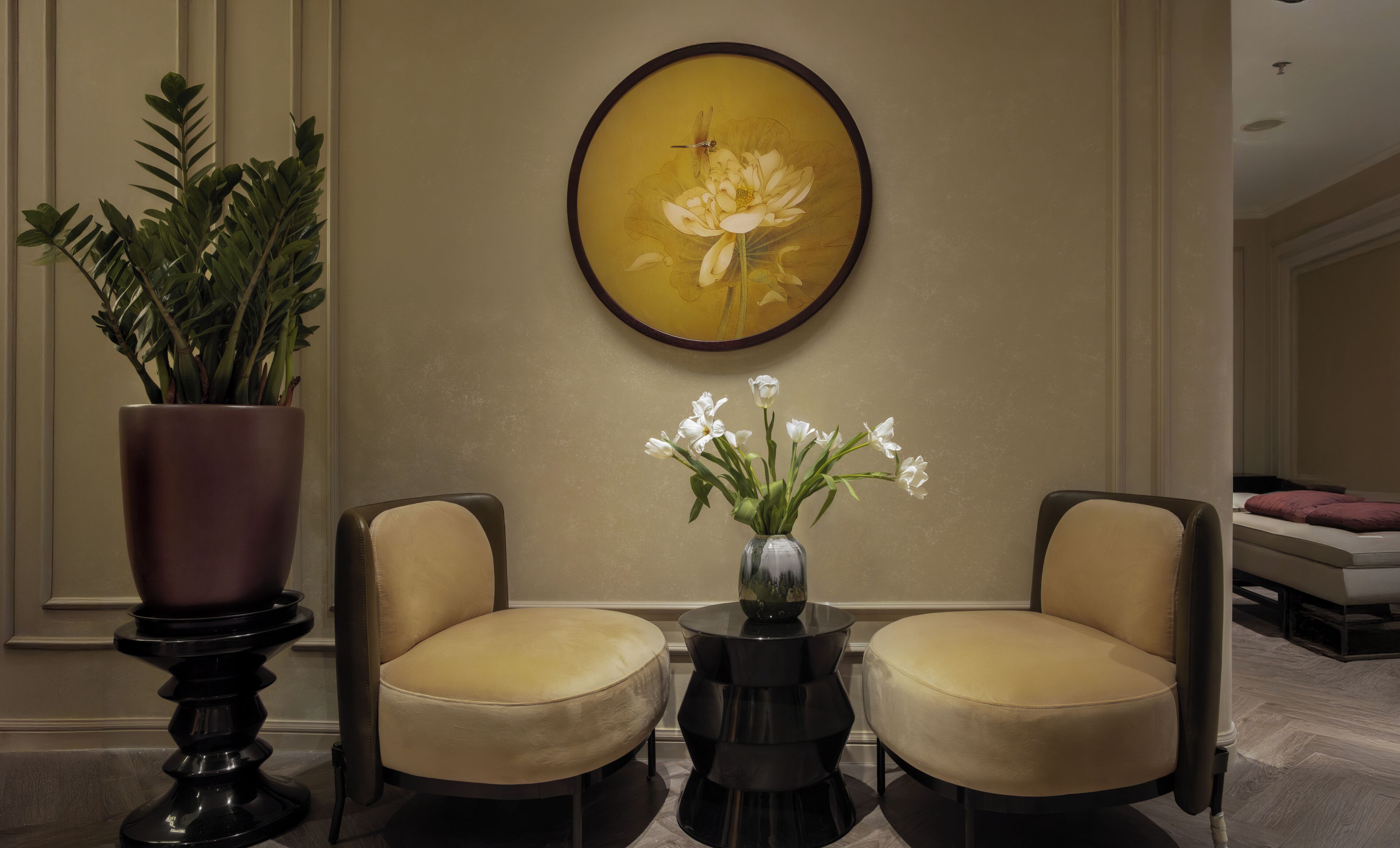 A lobby with two armchairs, a round table with a vase of flowers, and a large framed painting on the wall.