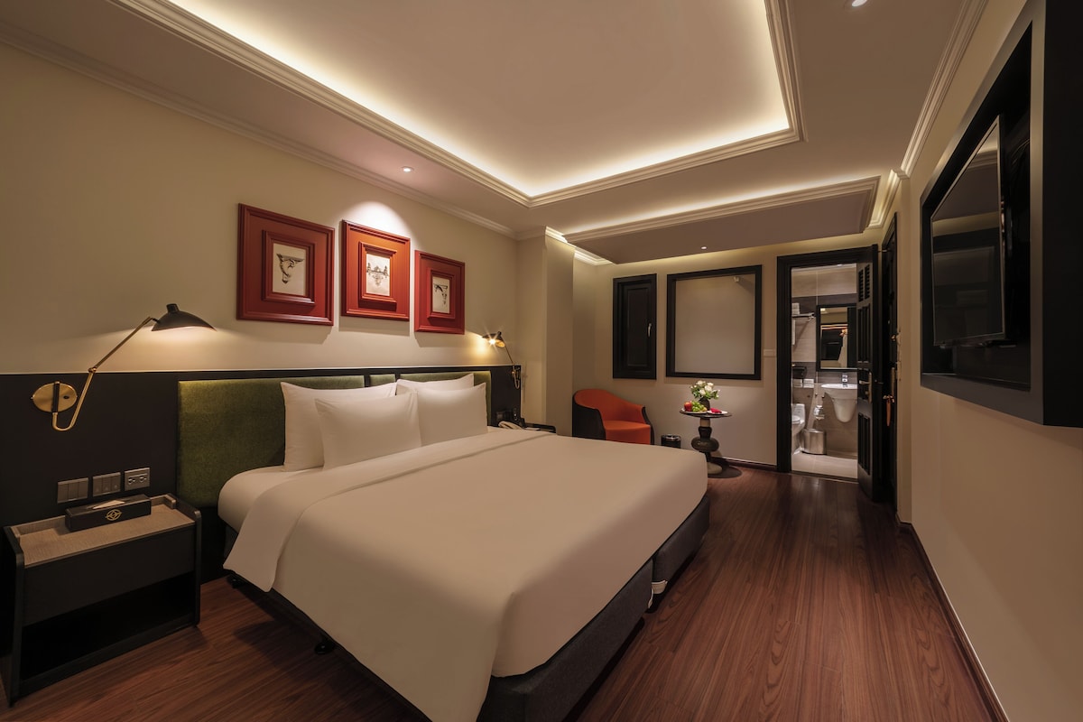 Habitación de hotel con una cama grande, una mesita de noche, un escritorio, una silla y un baño visible a través de una puerta abierta.