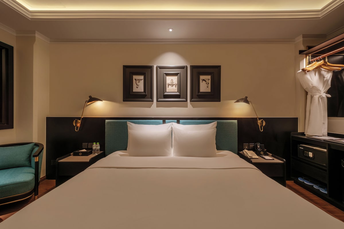 A hotel room with a large bed, two bedside tables, a nightstand, a chair, a wardrobe, and framed artwork on the wall.