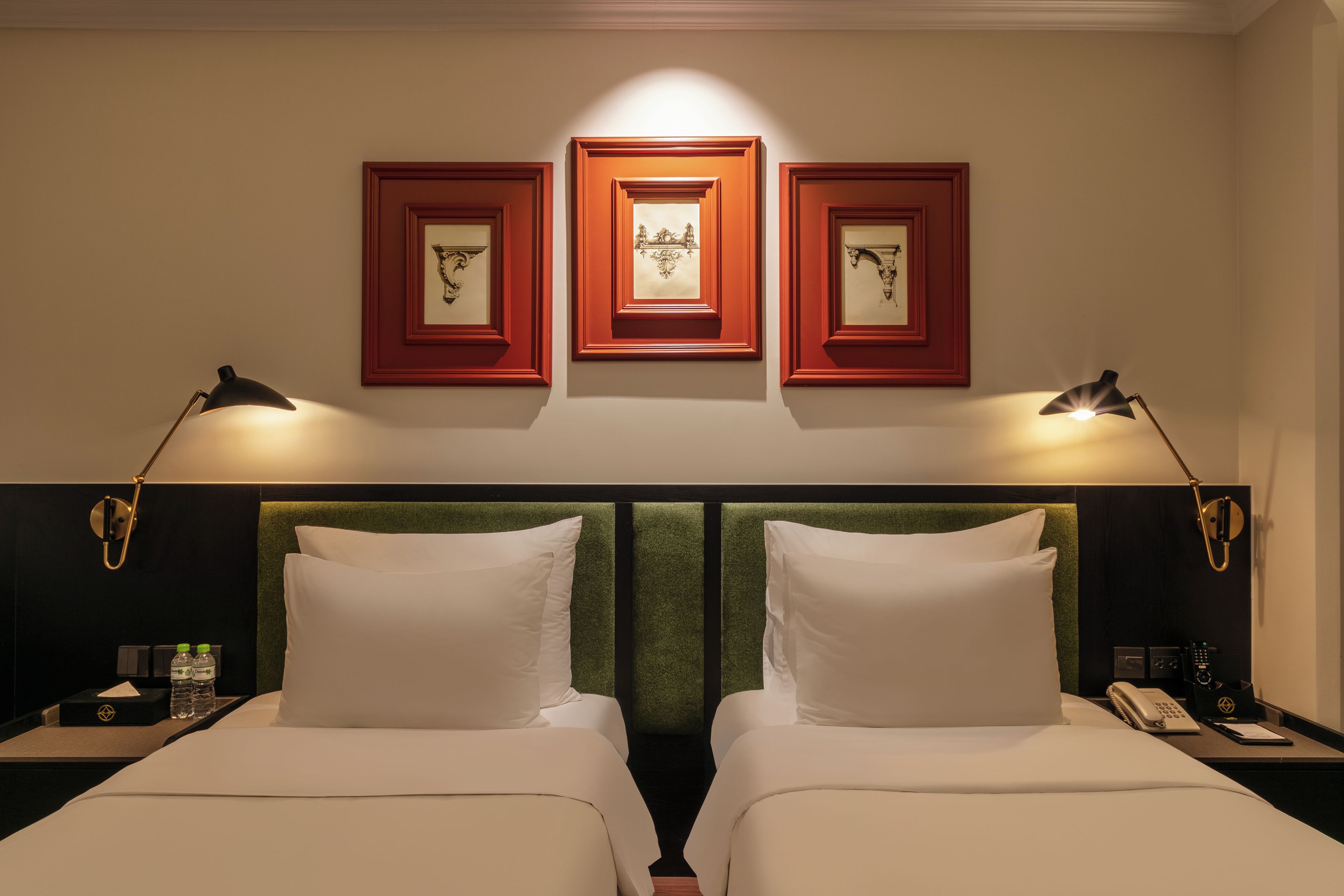 Een hotelkamer met twee bedden, een hoofdbord, nachtkastjes, lampen, ingelijste kunstwerken en een telefoon.
