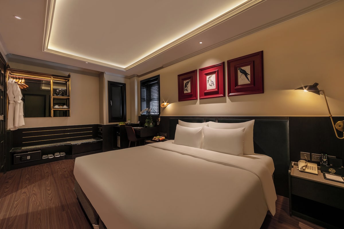 Una habitación de hotel con una cama grande, un escritorio con silla, un armario y un espejo. Hay tres cuadros en la pared.