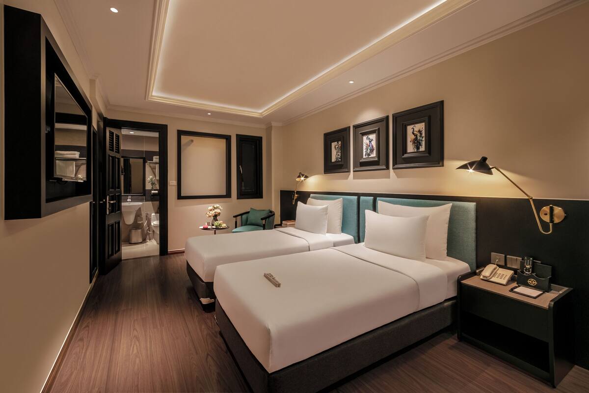 A hotel room with two beds, a desk, a chair, a nightstand, a lamp, and framed artwork on the wall.