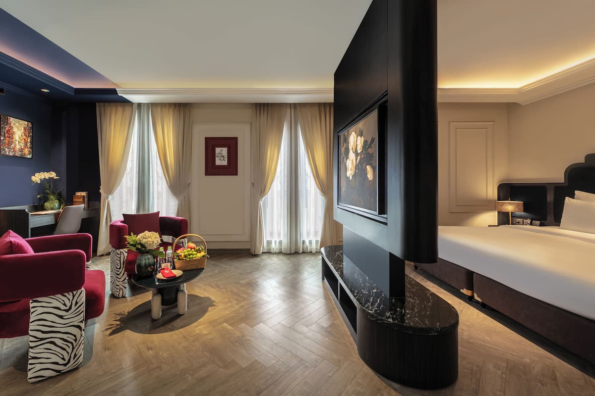 A hotel room with a bed, a TV, a seating area with chairs, a coffee table, and a vase of flowers.