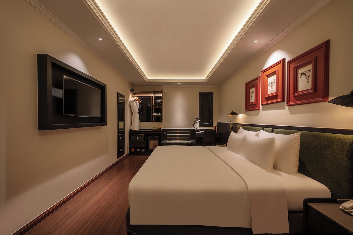 A hotel room with a large bed, a desk with a chair, a wardrobe, a TV mounted on the wall, and framed pictures on the wall.