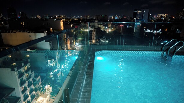 Una piscina en la azotea con agua cristalina y vistas nocturnas de la ciudad, rodeada de barandillas de vidrio.