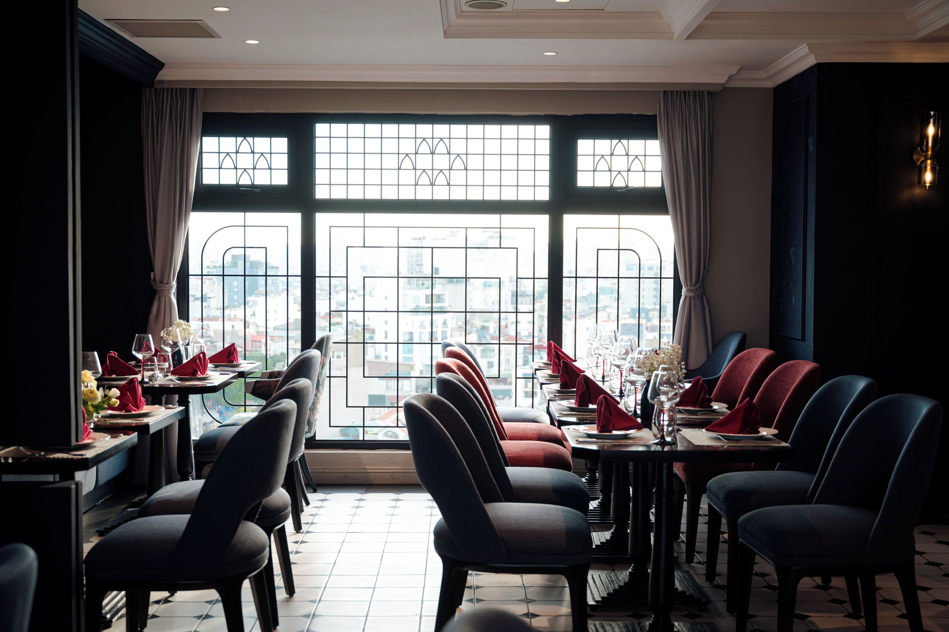 A restaurant with a view of the city through large windows, tables set with red napkins, and modern chairs.