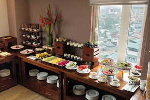 A buffet setup with various food items and a view of buildings through a window. - Champton Hanoi Hotel (Hanoi)