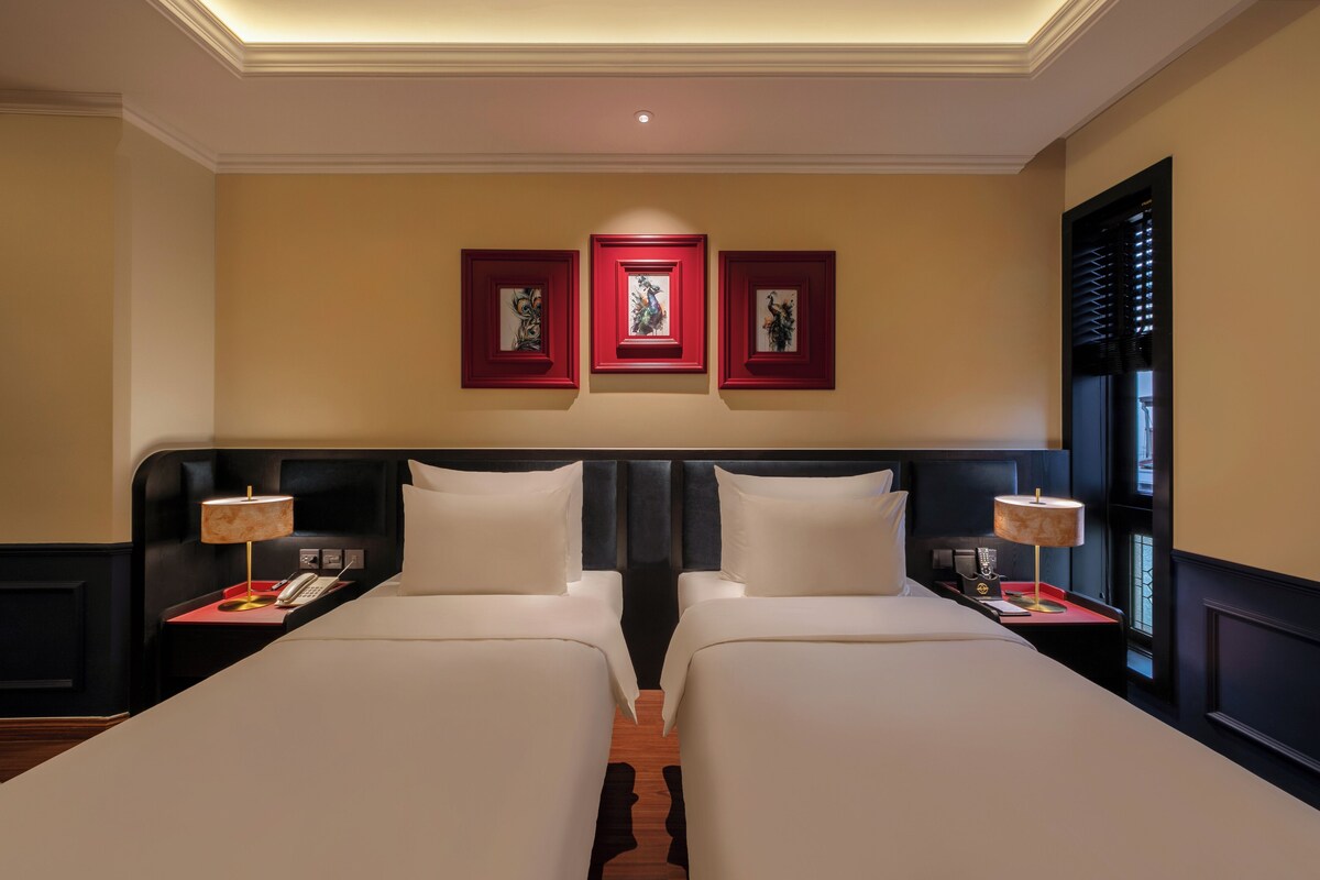 A hotel room with two beds, a dark headboard, a red-framed artwork on the wall, and a window with blinds.