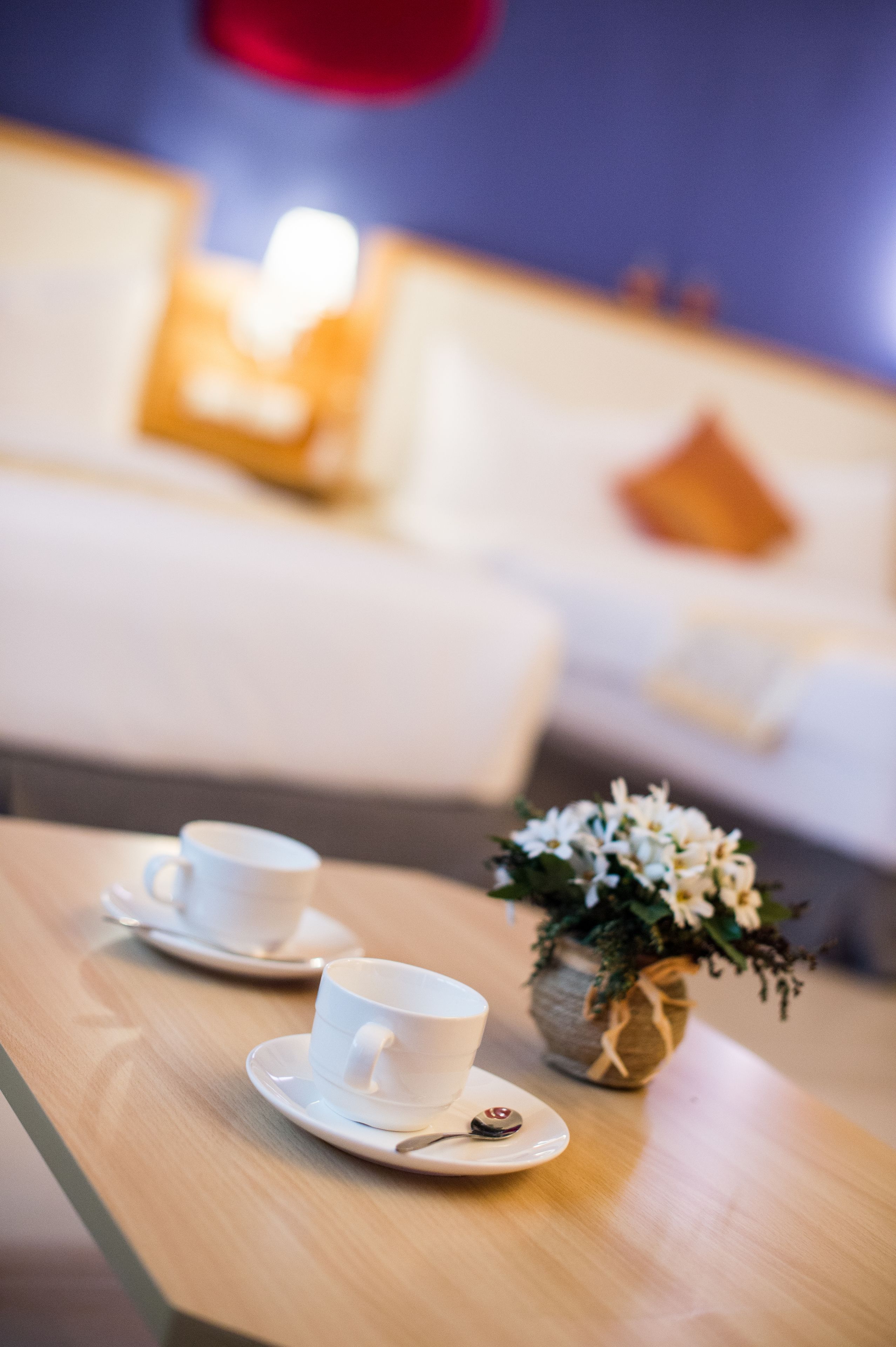 A hotel room with a wooden table, two white cups on saucers, a small vase with white flowers, and a blurred background of a bed and pillows.